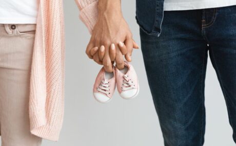 Cuplu de tineri, ea îmbrăcată cu un pulover roz-somon, tricou alb și jeanși bej, iar el cu cu tricou alb și cămașă și pantaloni de jeans, care se țin de mână și țin o pereche de pantofi de bebeluș, roz, ilustrând cum să rămâi cât mai repede însărcinată