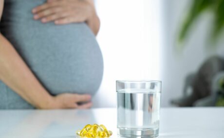 Femeie însărcinată, îmbrăcată într-o rochie mulată, gri, care își ține burta cu mâinile, având în față, pe masă, un pahar cu apă și câteva pastile galbene de vitamina D în sarcină