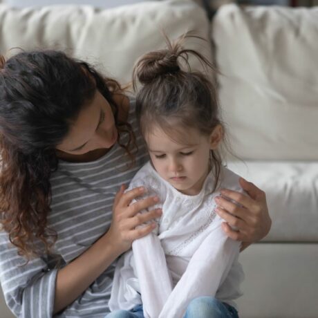 Mamă, îmbrăcată cu bluză gri cu dungi albe, stă așezată, în spatele fiicei ei, care are o bluză albă și jeanși care e tristă și își tine mâinile întinse în față, iar mama îi cuprinde umerii și se uită blând la ea, ilustrând anxietatea la copii
